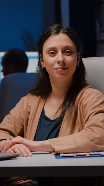 Portret uśmiechniętej bizneswoman patrzącej w kamerę siedząc przy biurku w startupowym biurze biznesowym późno w nocy