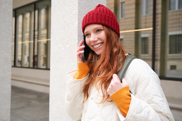 Bezpłatne zdjęcie portret uśmiechniętej imbirowej dziewczyny z czerwonym kapeluszem stoi na ulicy z plecakiem dzwoni do kogoś przez telefon a