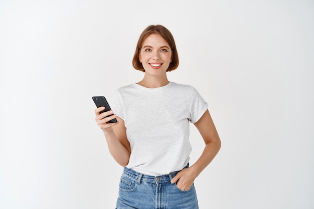Portret uśmiechniętej kobiety korzystającej ze smartfona, rozmawiającej w mediach społecznościowych, stojącej z telefonem komórkowym na tle białej ściany