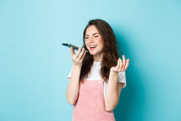 Portret uśmiechniętej kobiety trzymającej telefon w pobliżu ust i rozmawiającej, używającej tłumacza aplikacji na smartfonie lub nagrywającej wiadomość głosową, rozmawiającej przez zestaw głośnomówiący, stojącej na niebieskim tle