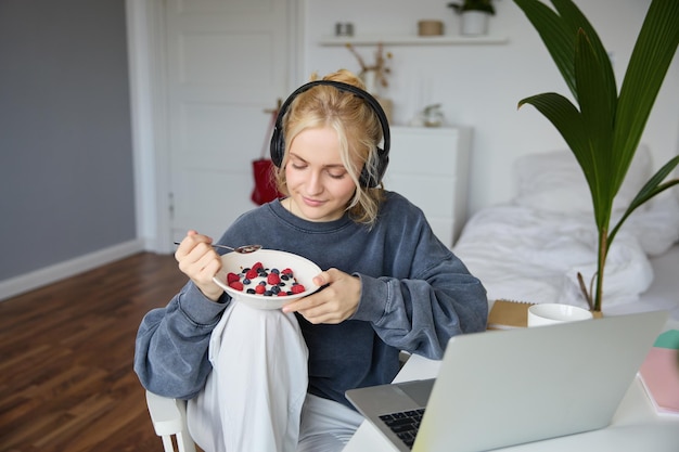 Bezpłatne zdjęcie portret uśmiechniętej młodej blondynki w słuchawkach siedzącej w pokoju oglądającej film na laptopie jedzącej