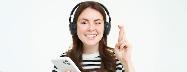 Bezpłatne zdjęcie portret uśmiechniętej modelki używającej słuchawek i smartfona trzymając palce skrzyżowane życzy sobie
