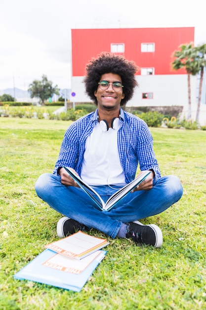 Portret uśmiechnięty afrykański męski uczeń siedzi na zielonej trawy mienia książce w ręce