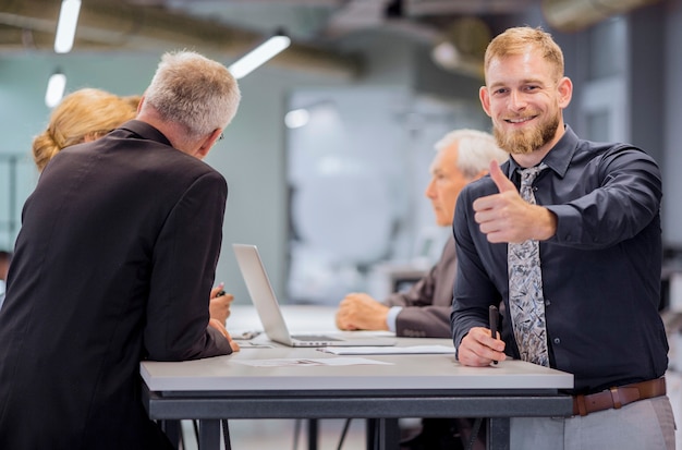 Bezpłatne zdjęcie portret uśmiechnięty biznesmen pokazuje kciuk up podpisuje podczas gdy drużynowy dyskutować w tle