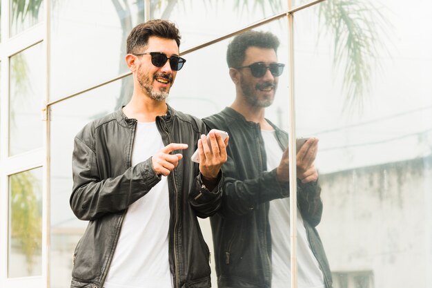 Portret uśmiechnięty elegancki mężczyzna jest ubranym okulary przeciwsłonecznych używać telefon komórkowego