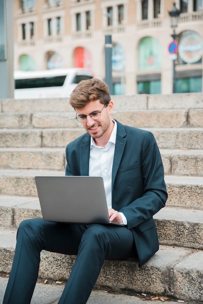 Portret uśmiechnięty młody biznesmena obsiadanie na schody używać laptop