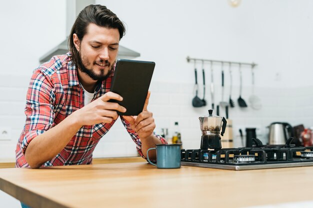 Portret uśmiechnięty młody człowiek opiera na drewnianym kuchennym kontuarze patrzeje mądrze telefon