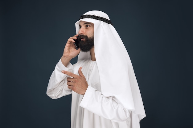 Portret w połowie długości arabski saudyjski mężczyzna na ciemnoniebieskiej ścianie studio. Model mężczyzna za pomocą smartfona, nawiązywanie połączenia. Pojęcie biznesu, finanse, wyraz twarzy, ludzkie emocje, technologie.