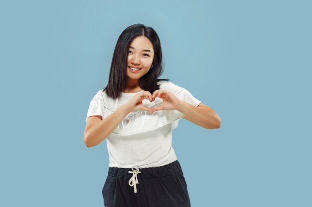 Portret w połowie długości koreański młodej kobiety na niebieskiej przestrzeni. Modelka w białej koszuli. Pokazuje znak serca.