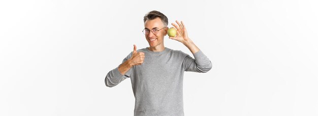 Bezpłatne zdjęcie portret wesołego, atrakcyjnego mężczyzny w średnim wieku, który czuje się zdrowo, pokazując kciuk do góry i zielone jabłko stojące nad białym tłem