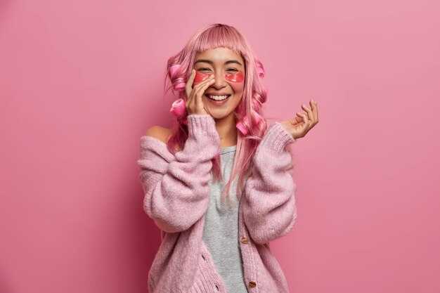 Portret wesołej Azjatki używa hydrożelowych plastrów o działaniu przeciwzmarszczkowym, nosi lokówki na różowych włosach, szczerze się uśmiecha, nosi swobodny sweter