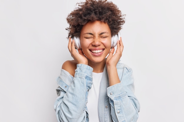 Portret wesołej kobiety z włosami afro trzyma ręce na słuchawkach stereo, ma zamknięte oczy, uśmiecha się szeroko, pokazuje białe zęby, cieszy się przyjemną muzyką na białym tle nad białą ścianą