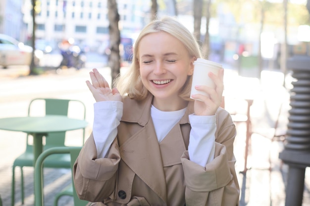 Bezpłatne zdjęcie portret wesołej młodej blondynki, która spędza czas w kawiarni na świeżym powietrzu, pijąc kawę i