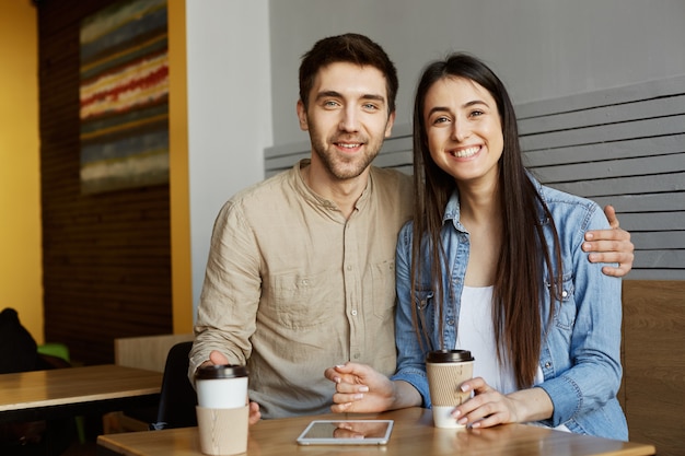 Portret wesołych perspektywicznych startupowców łączy się z ciemnymi włosami w przypadkowych ubraniach, siedzi w kawiarni, uśmiecha się jasno, pije kawę. i przytulanie.