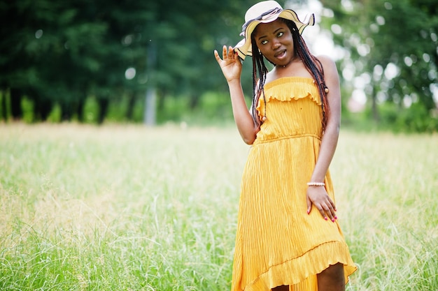 Portret wspaniałej Afroamerykanki w wieku 20 lat w żółtej sukience i letnim kapeluszu pozuje na zielonej trawie w parku