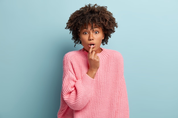 Portret wstrząśniętej Afroamerykanki sapie ze zdumienia, trzyma palec na ustach, oniemiała, słucha czegoś z zatroskaną miną, nosi różowy sweter, modelki w domu. Koncepcja OMG