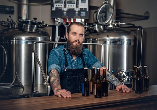 Portret wytatuowanego, brodatego męskiego producenta hipstera prezentującego piwo w minibrowarze.