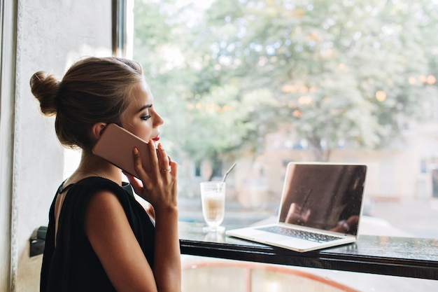 Bezpłatne zdjęcie portret z boku ładna kobieta w czarnej sukni w stołówce. mówi przez telefon, patrząc na laptopa.