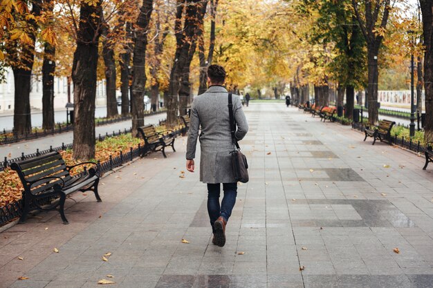 Portret z tyłu młodego stylowego faceta spacerującego w parku miejskim w płaszczu z torbą, patrząc na kolorowe drzewa