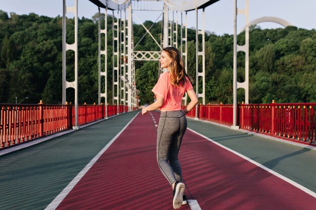 Portret z tyłu radosna dziewczyna biegnie rano i podziwiając widoki przyrody. Plenerowe zdjęcie inspirowanej kobiety wykonującej ćwiczenia.