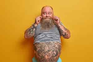 Bezpłatne zdjęcie portret zabawnego brodatego mężczyzny zwija wąsy, ma wytatuowane ramiona i brzuch, ubrany w niewymiarową koszulkę bez rękawów w paski, ma problem z otyłością i nadwagą, odizolowany na żółtej ścianie