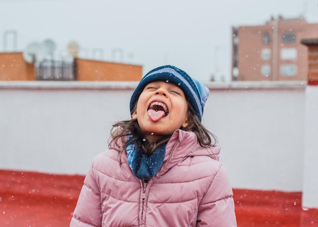 Portret zabawnej, słodkiej dziewczyny w ciepłym zimowym płaszczu i czapce łapiącej płatki śniegu na języku