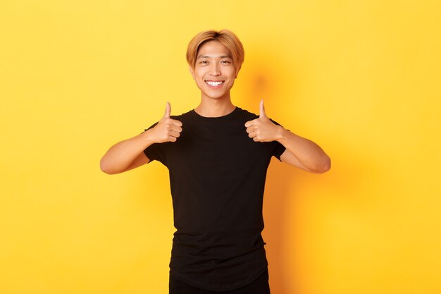 Portret zadowolony przystojny azjatycki student pokazuje aprobatę, stojąc na żółtej ścianie