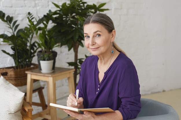 Portret zajętej, nowoczesnej 50-letniej kobiety w średnim wieku w fioletowej koszuli, piszącej w zeszycie, planującej, patrzącej z pozytywnym, przyjaznym uśmiechem, siedzącej na krześle, otoczonej zielonymi roślinami