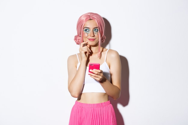 Bezpłatne zdjęcie portret zamyślonej dziewczyny ma ciekawy pomysł, odwracając wzrok i myśląc trzymając smartfona, nosząc różową perukę na imprezę, świętując wakacje.