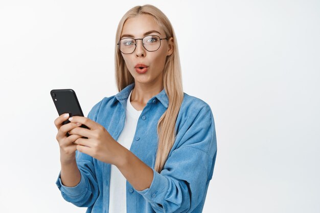 Portret zaskoczonej blond dziewczyny w okularach trzymającej smartfona i patrzącej zaintrygowanej na aparat stojący na białym tle