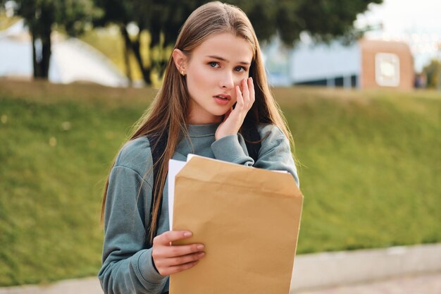 Portret zdenerwowanej przypadkowej studentki otwierającej kopertę z wynikami egzaminów, niestety patrzącej w kamerę w parku miejskim