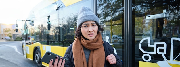 Portret zdezorientowanej Azjatki stojącej na przystanku autobusowym, trzymającej telefon komórkowy i wyglądającej na zszokowaną i zdenerwowaną