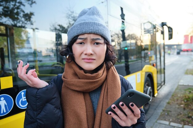 Portret zdezorientowanej azjatyckiej dziewczyny stojącej na przystanku autobusowym z telefonem komórkowym, wyglądającej na zszokowaną i zdenerwowaną