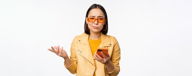 Portret zdezorientowanej azjatyckiej dziewczyny w okularach przeciwsłonecznych, trzymającej smartfona za pomocą telefonu komórkowego i patrzącego na zdziwionego, stojącego nieświadomie na białym tle