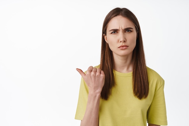 Portret zdezorientowanej brunetki wskazującej w lewo, mrużącej oczy i marszczącej brwi, zdziwiona, pokazująca coś dziwnego, zadająca pytanie, co to za gest, białe tło