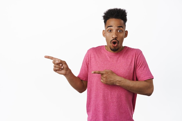Bezpłatne zdjęcie portret zdziwionego afroamerykanina wzdycha, mówi wow i gapi się zdumiony, wskazując palcami zostawionymi na logo zniżki wyprzedaży, pokazując reklamę, stojący w różowej koszulce na białym tle.