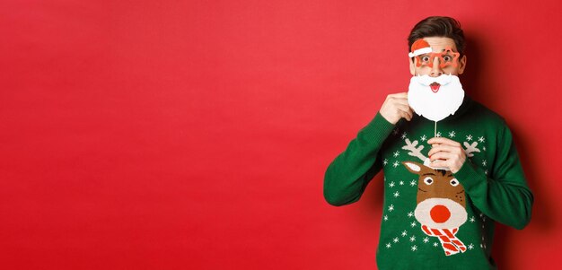 Portret zdziwionego mężczyzny w zielonym świątecznym swetrze trzymającym śmieszną maskę świętego mikołaja świętującego nowy ...