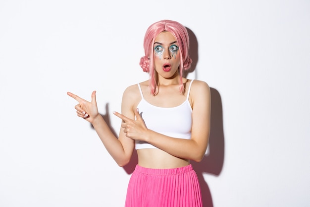 Portret zdziwionej modelki w różowej peruce anime i makijażu imprezowym, wskazując palcami i patrząc w lewo z pod wrażeniem wyrazu twarzy, stojąc na białym tle