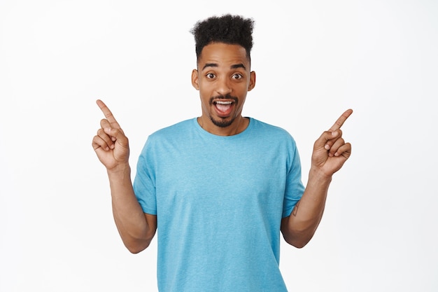 Portret Zdziwiony African American Man Wskazując Palcami W Bok, Pokazując Lewy I Prawy Produkt, Dwa Rabaty Sprzedaży, Stojąc W Niebieskim T-shirt Na Białym Tle.