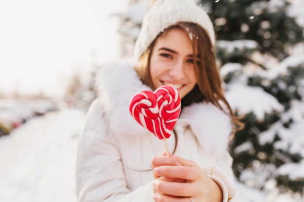 Portret zima młoda kobieta trzyma różowe serce lollypop, chłodzenie na ulicy pełnej śniegu w słoneczny poranek. Biała czapka z dzianiny, uśmiechnięta.