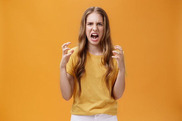 Portret zirytowanej uroczej europejskiej kobiety ściskającej dłonie w pięści z gniewem marszczącym brwi i robiące nienawistną twarz stojącą zirytowaną i wkurzoną na pomarańczowym tle z wściekłym spojrzeniem. Koncepcja emocji