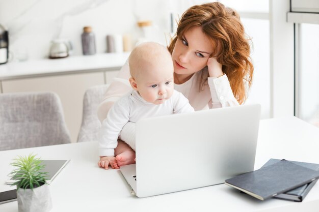 Portret zmęczonej kobiety biznesu siedzącej przy stole i pracującej na laptopie, trzymając w pobliżu swoje małe dziecko