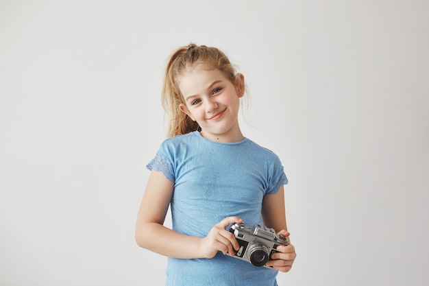 Portreta o przystojny blond dziecko ono uśmiecha się w błękitnej koszulce, stojący z aparatem fotograficznym w rękach pozuje dla szkolnego albumu.