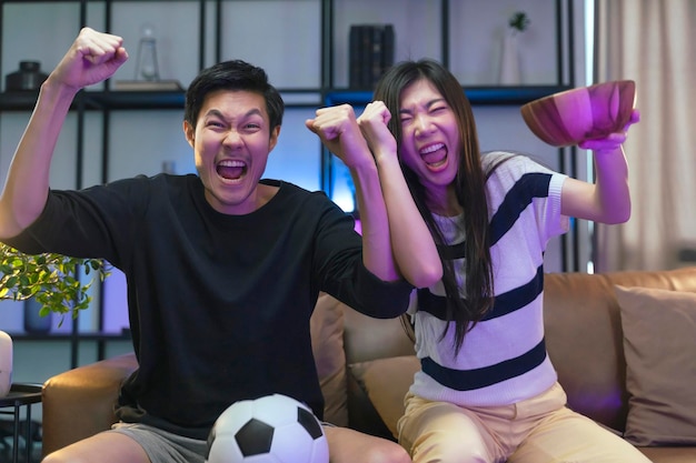Poślubić dorosłego Azjatę oglądającego telewizję w domu azjatycka para wiwatująca sportowa rywalizacja razem ze śmiechem uśmiech zwycięstwo na kanapie kanapie w salonie izolacja domu