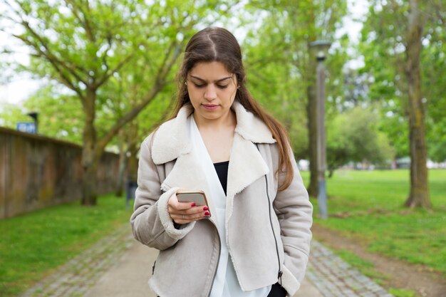 Poważna ładna młoda kobieta używa smartphone w parku