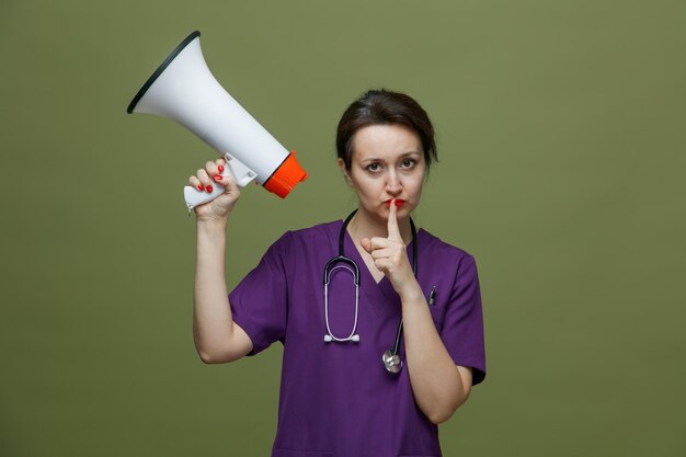 Poważna Lekarka W średnim Wieku Nosząca Mundur I Stetoskop Na Szyi, Trzymająca Głośnika Patrzącego Na Kamerę Pokazującą Gest Ciszy Na Oliwkowym Tle