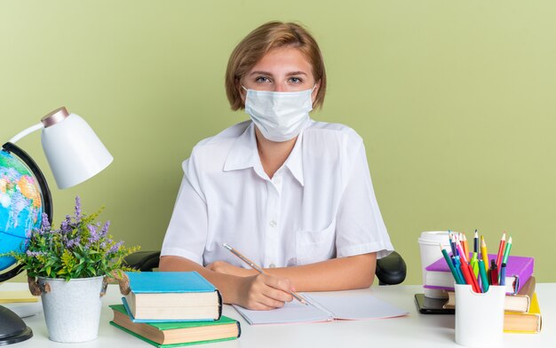 Poważna młoda blond studentka w masce ochronnej siedzi przy biurku z szkolnymi narzędziami trzymając ołówek, patrząc na kamerę odizolowaną na oliwkowozielonej ścianie