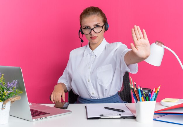 Poważna młoda blondynka call center dziewczyna ubrana w zestaw słuchawkowy i okulary, siedząca przy biurku z narzędziami do pracy, wykonująca gest zatrzymania