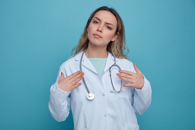 Poważna młoda kobieta lekarz ubrana w szlafrok medyczny i stetoskop wokół szyi, wskazując na siebie rękami