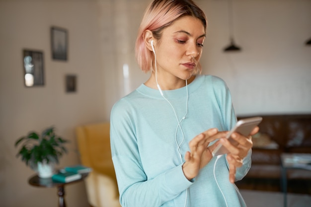 Bezpłatne zdjęcie poważna młoda kobieta z kolczykiem na twarzy, pracująca w oddali przy użyciu telefonu komórkowego, słuchająca muzyki w słuchawkach